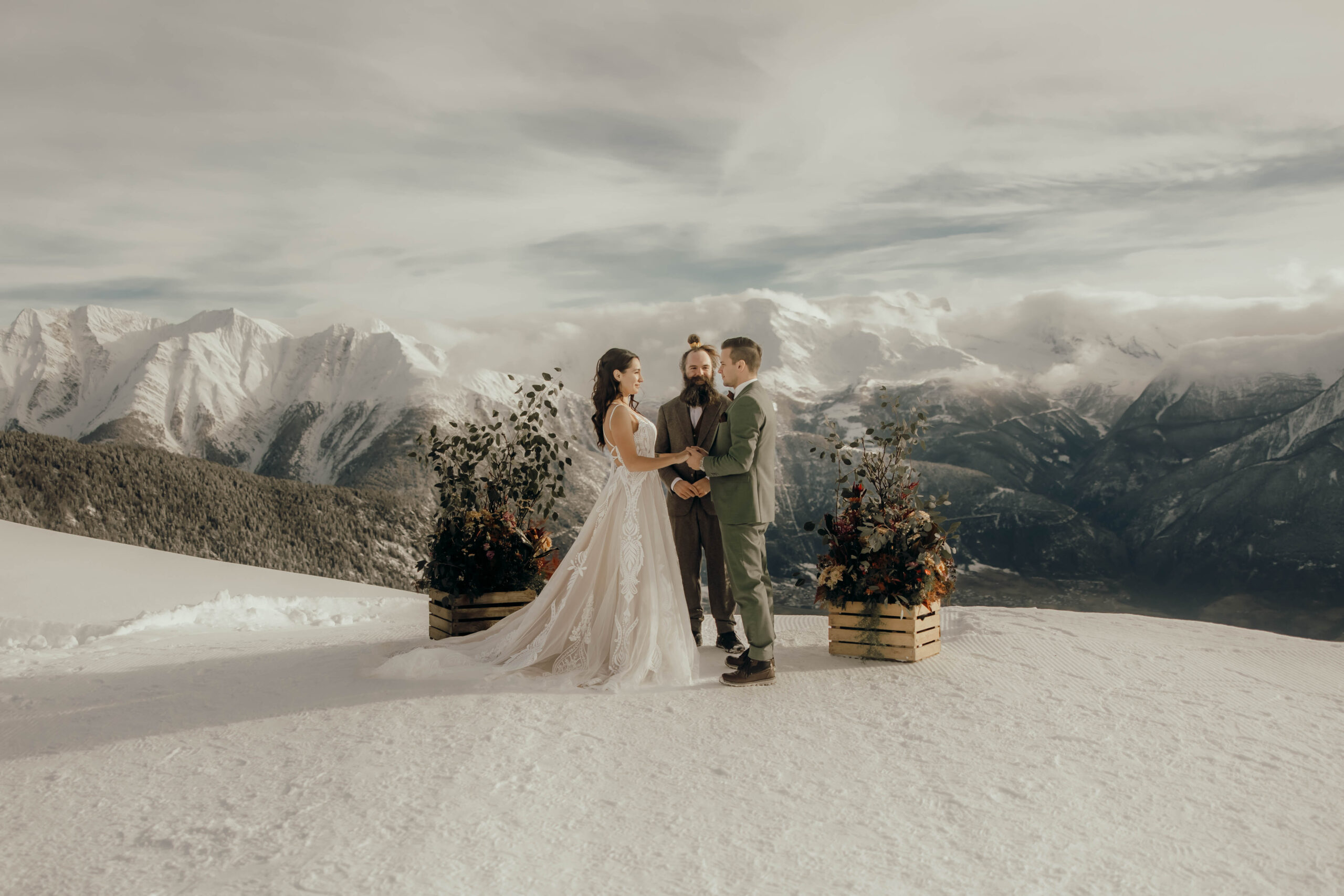 Ein Paar heiratet auf einem schneebedeckten Berggipfel in Belalp, mit einem malerischen Panoramablick auf schneebedeckte Berge im Hintergrund. In Begleitung eines Geistlichen und umgeben von Blumenarrangements in Holzkisten haben sie sich für ihren besonderen Tag das Wallis ausgesucht. Der Himmel ist teilweise bewölkt.