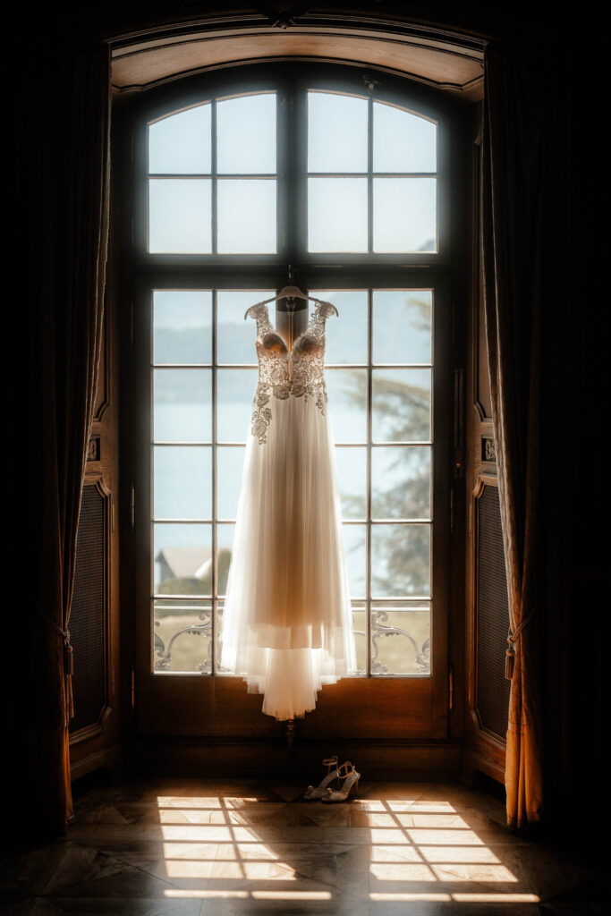 Ein mit aufwendigen Spitzendetails verziertes Brautkleid hängt vor einem großen, gewölbten Fenster mit Blick auf eine ruhige Außenszene. Sanftes Sonnenlicht fällt hindurch und erhellt das Kleid. Ein Paar Schuhe liegt auf dem Boden darunter, perfekt eingefangen vom Hochzeitsfotografen in meggen.