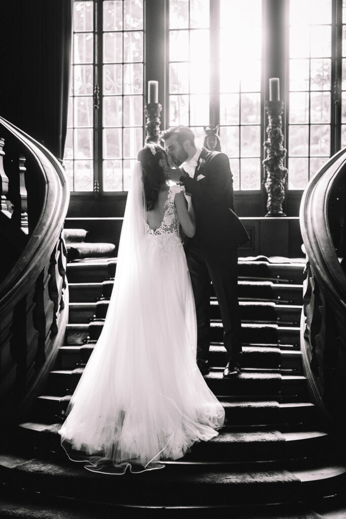 Brautpaar kuesst sich oben auf einer Treppe in einem prächtigen Gebäude mit großen Fenstern und Holzgeländern, wunderschön eingefangen von ihrem Hochzeitsfotografen.