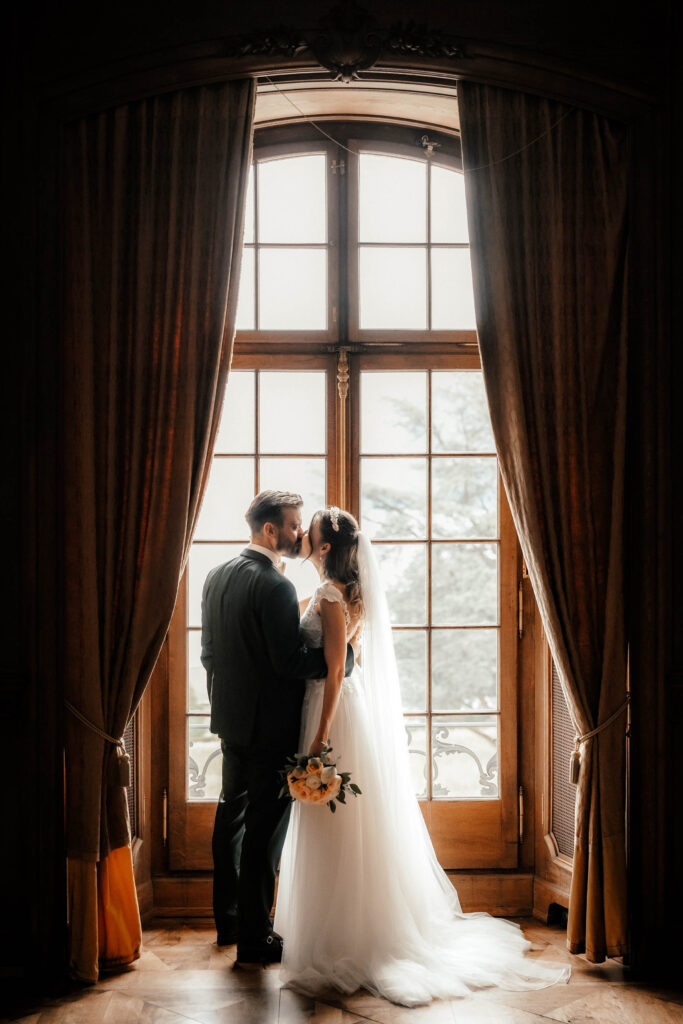 Brautpaar kuesst sich romantisch vor einem hohen, eleganten Fenster, das von langen Vorhängen eingerahmt wird. Die Braut trägt ein fließendes weißes Kleid und einen Schleier und hält einen Blumenstrauß in der Hand. Der Bräutigam trägt einen dunklen Anzug. Natürliches Licht strömt herein und wirft einen sanften Schein, der von ihrem Hochzeitsfotografen in Meggen perfekt eingefangen wurde.